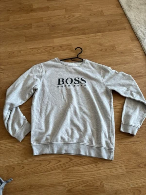 Grå sweatshirt från Hugo Boss - Snygg grå sweatshirt från Hugo Boss med svart logga på bröstet. Tröjan har rund halsringning, långa ärmar och ribbade muddar vid ärmslut och nederkant. Perfekt för en avslappnad och stilren look.