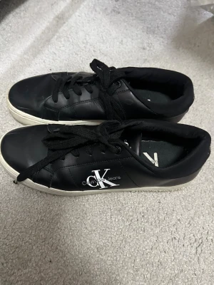 Svarta Calvin Klein Jeans sneakers - Säljer ett par svarta sneakers från Calvin Klein Jeans med vit logga på sidan. Skorna har klassisk låg modell, svarta skosnören och vit sula. Materialet är syntetläder och insidan är mjukt fodrad för extra komfort.