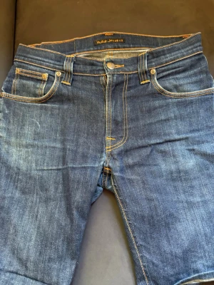 Mörkblå jeans från Nudie Jeans - Säljer ett par klassiska mörkblå jeans från Nudie Jeans med kontrastsömmar och femficksmodell. Jeansen har en rak passform och detaljerade bakfickor med Nudie Jeans karakteristiska brodyr. Materialet är slitstark denim i bomull.