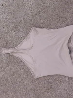 Beige body med breda axelband - Säljer en enkel och stilren beige body med breda axelband och rundad halsringning. Bodyn har knäppning i grenen och är tillverkad i ett mjukt, stretchigt material som sitter skönt mot kroppen. Perfekt att styla till olika outfits. Aldrig använd 