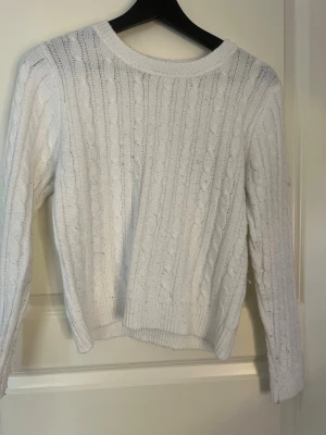 Vit kabelstickad tröja - Kabelstickad vit tröja med rund hals och långa ärmar. Tröjan har ett klassiskt flätmönster och ribbade muddar vid ärmslut och nederkant. Perfekt för lager på lager och en clean look.