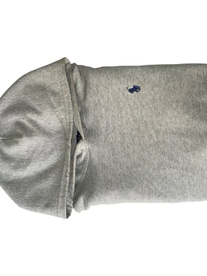 Grå hoodie från Polo Ralph Lauren - Snygg grå hoodie från Polo Ralph Lauren med klassisk blå logga broderad på bröstet. Hoodien har huva och ribbade muddar vid ärmslut och nederkant. Tillverkad i mjukt bomullsmaterial som är skönt att bära. Perfekt för en avslappnad och stilren look.