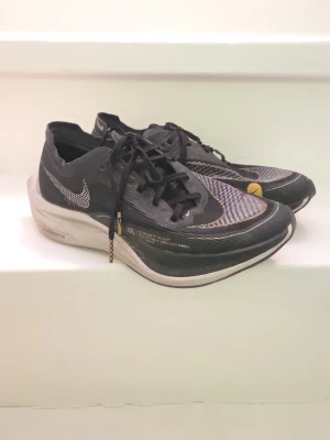 Nike ZoomX Vaporfly 2% löparskor - Svarta Nike ZoomX Vaporfly 2% löparskor med vit sula och detaljer i vitt och guld. Skorna har en sportig design med snörning och synlig Nike-logga på sidan. Ovandelen är i meshmaterial för bra ventilation och lätt känsla. Carbonplatta vilket gör att du springer snabbare med lägre ansträngning. 