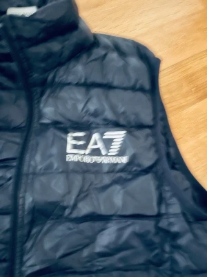 Svart dunväst från Emporio Armani EA7 - Snygg svart dunväst från Emporio Armani EA7 med quiltad design och dragkedja framtill. Västen har hög krage och logga på bröstet. Perfekt för lager-på-lager och streetstyle. Använd lite . Ny pris 1300 kr.