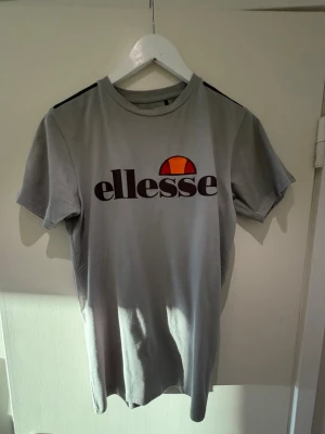 Grå t-shirt från Ellesse - Snygg grå t-shirt från Ellesse med stor logga framtill i svart och orange. Klassisk rund halsringning och korta ärmar. Perfekt för en avslappnad och sportig stil.