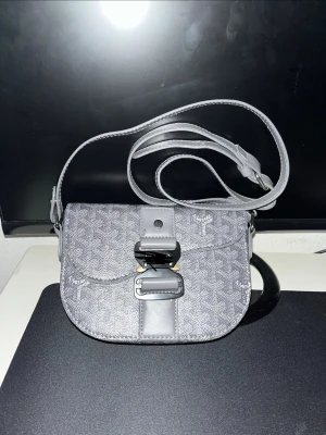 Grå axelväska  - Snygg grå axelväska  med justerbar axelrem och klassiskt monogrammönster. Väskan har rundad form, lock med spänne och detaljer i läder. Perfekt för dig som gillar stilrena accessoarer med logga.