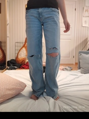 Ljusblå slitna jeans med knappar - Ljusblå jeans med raka ben och stora slitningar över knäna. Dekorerade med vita och guldiga knappar på bakfickan för en unik look. Klassisk femficksmodell i denim, perfekt för dig som gillar streetstyle och detaljer.