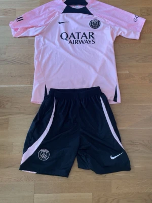PSG matchställ rosa/svart Nike - Säljer ett PSG matchställ från Nike med tröja i ljusrosa och svarta detaljer samt svarta shorts med rosa inslag. Tröjan har Qatar Airways-tryck och PSG-logga på bröstet. Shortsen har elastisk midja och PSG-logga. Perfekt för fotbollsträning eller streetstyle. Dragkedjan på en av fickorna i shortsen har fastnat i mitten av shortsen, annars är kläderna så gott som nya. Vid minsta lilla fråga, kontakta gärna. 