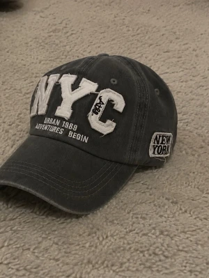 Grå NYC keps med brodyr - Säljer en grå keps med stora vita broderade bokstäver 'NYC' framtill och texten 'Urban 1989 Adventures Begin'. På sidan finns en patch med 'New York'. Kepsen har böjd skärm och justerbar rem bak. Perfekt för dig som gillar streetstyle.