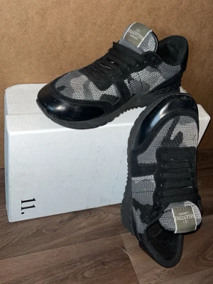 Valentino Garavani svarta sneakers - Snygga svarta sneakers från Valentino Garavani med grått och svart kamouflagemönster. Skorna har svarta snören, mesh- och läderdetaljer samt en robust sula. Perfekta för dig som vill ha en unik och trendig look.