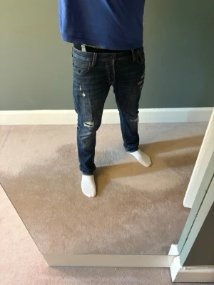 Slim fit jeans med slitningar - Säljer ett par mörkblå jeans med slitningar och hål på både lår och knän. Jeansen har en smal passform och klassisk femficksdesign. Perfekta för dig som gillar en avslappnad och trendig stil. 