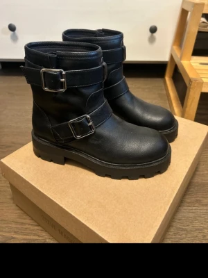 Svarta bikerboots  - Tuffa svarta bikerboots från Steve Madden i läder med grov sula och två breda spännen i silver. Stövlarna har rund tå och en cool, chunky look som passar perfekt till jeans eller kjol. Snygga detaljer och robust design för en edgy stil. Använda 2 gånger.