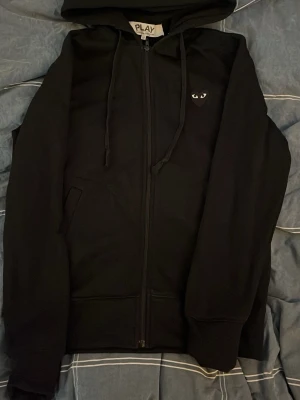 Svart hoodie Comme des Garçons Play - Svart hoodie med dragkedja från Comme des Garçons Play. Klassisk design med huva, dragsnören och fickor. Liten ikonisk hjärtlapp med ögon på bröstet. Perfekt för en clean och stilren look.