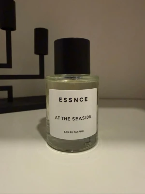 ESSNCE At The Seaside parfym -  ESSNCE med doften At The Seaside. Flaskan är genomskinlig med en svart cylinderformad kork och vit etikett med svart text. Eau de Parfum-varianten ger en fräsch och modern känsla.