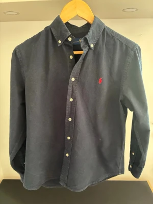 Mörkblå skjorta från Ralph Lauren - Säljer en klassisk mörkblå skjorta från Ralph Lauren med button-down krage och vita knappar. Skjortan har långärmad design och ikonisk röd logobrodyr på bröstet. Perfekt för dig som gillar stilrena plagg med premiumkänsla. Passar någon som är ca 160 cm