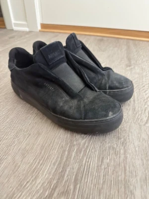 Svarta sneakers från Axel Arigato - Stilrena svarta sneakers från Axel Arigato i mocka med minimalistisk design och bred elastisk rem över vristen istället för klassiska snören. Sulan är svart och loggan syns diskret på sidan. Perfekta för en clean och modern look.