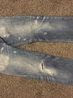 Blå slitna jeans med distressed detaljer - Säljer ett par blå jeans med tydliga slitningar och distressed detaljer på både fram- och baksida. Jeansen har en tvättad look med ljusare partier och flera små hål och revor för en trendig, avslappnad stil. Perfekta för dig som gillar streetwear och en edgy vibe.