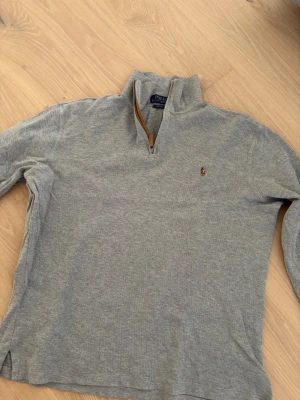Grå halvzip från Polo Ralph Lauren - Snygg grå tröja från Polo Ralph Lauren med halv dragkedja och ståkrage. Klassisk broderad logga på bröstet och ribbade muddar. Perfekt till jeans eller chinos för en clean och stilren look.
