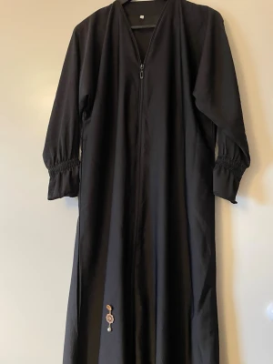 Svart långklänning med dragkedja / Abaya - Säljer en svart långklänning med hel dragkedja framtill och långa ärmar med smockdetalj vid ärmsluten. Klänningen har en enkel, rak passform och en liten dekorativ detalj längst ner. Perfekt för dig som gillar stilrena plagg. Passar 155-163cm. One size fits all. Aldrig använd.