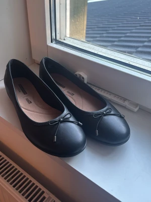 Svarta ballerinaskor med rosett - Stilrena svarta ballerinaskor från DinSko med rund tå och liten dekorativ rosett framtill. Skorna har en klassisk form och är tillverkade i ett slätt syntetmaterial med beige innersula för extra komfort.