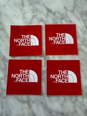 The North Face klistermärken röda - Fyra röda fyrkantiga klistermärken med The North Face-loggan i vitt. Perfekta att sätta på laptop, mobil eller vattenflaska för att ge din stil en extra touch. Tillverkade i plastmaterial med självhäftande baksida.