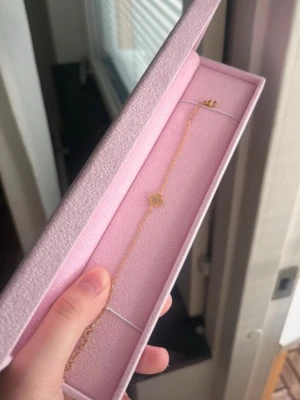 Guldfärgat armband med blomma HIMO - Ett tunt och elegant armband i guldfärg med en liten blomformad detalj i mitten. Armbandet har en fin kedja och kommer i en rosa ask. Perfekt för dig som gillar stilrena och söta accessoarer.