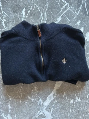 Mörkblå halvzip-tröja från Morris - Mörkblå stickad tröja från Morris med halv dragkedja och beige insida på kragen. Tröjan har en liten broderad lilja på bröstet och ribbstickade detaljer vid hals och ärmslut. Perfekt för lager-på-lager och snygg till jeans.