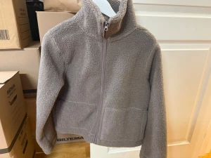 Grå/brun teddyjacka med dragkedja - Mysig grå/brun teddyjacka med hög krage och hel dragkedja framtill. Jackan har två sidofickor och är tillverkad i ett fluffigt, mjukt material som ger en avslappnad look. Perfekt för kyliga dagar när du vill vara både varm och stilren. Jackan är använd 1 gång men den känndes för liten så mycket bra skick!