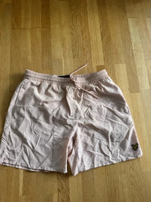 Ljusrosa badbyxor från Lyle & Scott - Snygga ljusrosa badbyxor från Lyle & Scott med elastisk midja och dragsko. Klassisk mesh-innerbyxa och gul logga på vänster ben. Perfekta för stranden eller poolen, tillverkade i snabbtorkande material.