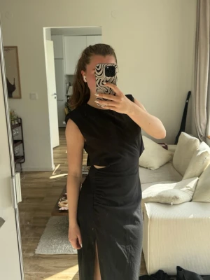 Svart långklänning med slits - Säljer en svart långklänning med ärmlös design och hög slits framtill. Klänningen har en enkel och stilren form med rund halsringning och diskret cut-out på sidan. Perfekt för dig som gillar minimalistisk stil och vill ha något elegant.