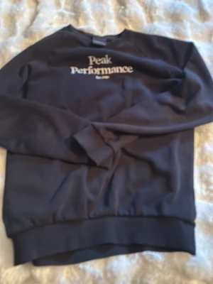 Svart sweatshirt Peak Performance - Klassisk svart sweatshirt från Peak Performance med vit logga på bröstet. Tröjan har rund halsringning, långa ärmar och ribbade muddar vid ärmslut och nederkant. Perfekt för en avslappnad och stilren look.