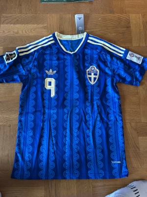 Blå Sverige fotbollströja Adidas - Blå fotbollströja från Adidas med Sveriges landslagsmärke och nummer 9 på bröstet. Tröjan har gul och vit krage, vita ränder på axlarna och detaljerade mönster i tyget. Perfekt för dig som vill stötta Sverige med stil.