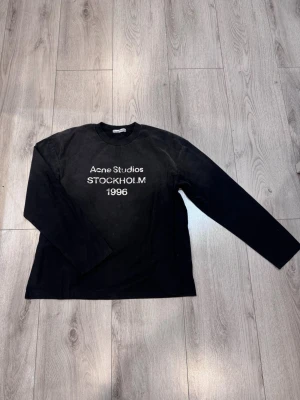 Svart långärmad tröja Acne Studios - Svart långärmad tröja från Acne Studios med vit text 'Acne Studios STOCKHOLM 1996' på bröstet. Rund halsringning och rak passform. Perfekt för en stilren och enkel look. Tröjan har en oversized passform så den kan även passa dig med storlek Medium