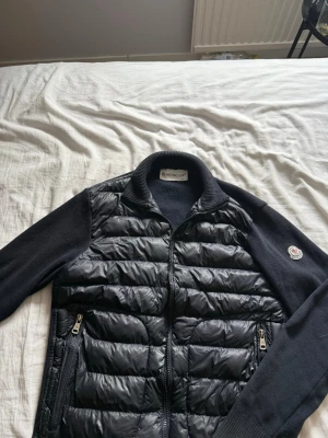 Svart cardiganjacka från Moncler - Snygg svart cardiganjacka från Moncler med quiltad front i glansigt material och stickade ärmar. Jackan har dragkedja framtill, två fickor med dragkedja och Moncler-logga på ärmen. Perfekt för lager-på-lager och streetstyle.