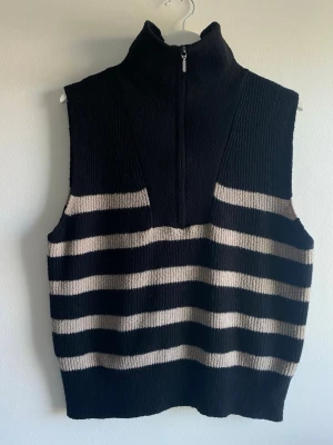 Randig stickad väst från Gina Tricot S - Snygg stickad väst från Gina Tricot med breda svarta och beige ränder. Västen har hög krage med half zip-dragkedja och är ärmlös. Perfekt att styla över en skjorta eller t-shirt för en trendig lager-på-lager-look.