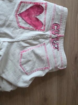 Vita Guess jeans med rosa detaljer - Unika vita jeans från Guess med raka ben och handmålade rosa detaljer. Bakfickorna har rosa kanter, ena fickan har ett stort rosa hjärta och loggan är målad i rosa. Jeansen har klassisk femficksmodell och är tillverkade i denim. Jeansen er highwaist och fran hm