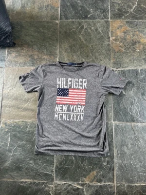 Grå Hilfiger t-shirt med USA-flagga - Snygg grå t-shirt från Tommy Hilfiger med tryck av amerikanska flaggan och texten 'HILFIGER NEW YORK MCMLXXXV' på bröstet. Klassisk rund hals och korta ärmar. Perfekt för dig som gillar streetwear och vill ha en casual look.
