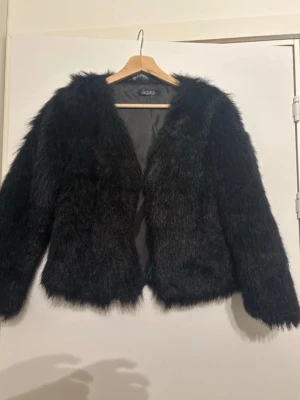 Svart fuskpälsjacka från Zizzi - Säljer en svart fuskpälsjacka från Zizzi med mjukt och fluffigt material. Jackan har en rak passform, långa ärmar och är helsvart med diskret glans. Perfekt för dig som vill ha en trendig och varm look under vintern.