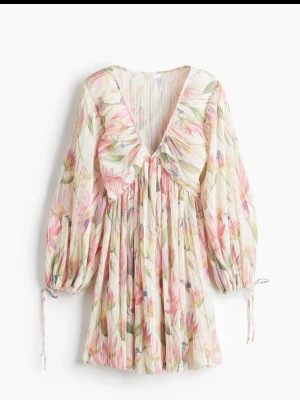 Blommig plisserad kortklänning H&M - Superfin kortklänning från H&M med plisserat tyg och blommigt mönster i rosa, grönt och vitt. Klänningen har v-ringning, volangdetalj framtill och långa ärmar med knyt vid ärmslut. Perfekt för vår och sommar!