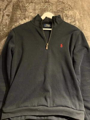 Mörkblå quarter zip från Polo Ralph Lauren - Mörkblå tröja med quarter zip från Polo Ralph Lauren. Klassisk design med röd broderad logga på bröstet och hög krage. Tillverkad i ett mjukt och bekvämt material, perfekt för lager-på-lager. Snygg och stilren för en avslappnad look. Storlek S förkortad