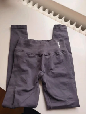 Grå seamless leggings från DFYNE - Säljer ett par grå seamless leggings från DFYNE med hög midja och ribbad linning. Materialet är stretchigt och följsamt, perfekt för träning eller chill. Diskret logga framtill i vitt och scrunch-detalj bak för extra passform.