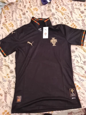 Portugal special edition fotbollströja Puma - Säljer en svart Portugal special edition fotbollströja från Puma med guldfärgade detaljer och emblem. Tröjan har ribbade muddar med gröna och röda ränder, ståkrage och är tillverkad i funktionsmaterialet Drycell. Perfekt för dig som älskar fotboll och samlartröjor.