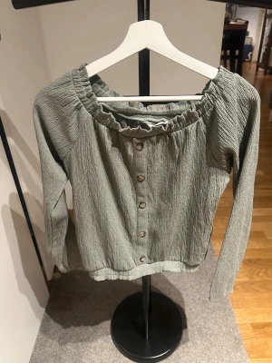 Grön crinkle-blus med knappar - Trendig grön blus i crinkle-material med långa ärmar och knappar framtill. Blusen har en bred resår vid halsringningen som ger en snygg rynkad effekt. Perfekt att styla med jeans eller kjol för en avslappnad look.