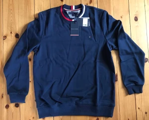 Marinblå sweatshirt Tommy Hilfiger - Snygg marinblå sweatshirt från Tommy Hilfiger med rund hals och ribbade muddar. Kragen har röd, vit och marinblå randdetalj och den klassiska lilla loggan broderad på bröstet. Perfekt för en stilren och sportig look.