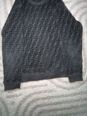 Svart Fendi sweatshirt med logomönster - Cool svart sweatshirt från Fendi med det klassiska FF-logomönstret över hela tröjan. Tröjan har rund halsringning, ribbade muddar och är tillverkad i ett mjukt material som känns skönt mot huden. Perfekt för dig som gillar streetwear och designerplagg.