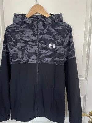Svart vindjacka från Under Armour - Snygg svart vindjacka från Under Armour med grått camomönster upptill och på huvan. Jackan har dragkedja framtill, två sidofickor och en vit logga på bröstet. Tillverkad i lätt och vindtätt material, perfekt för blåsiga dagar.