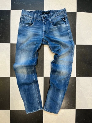 Replay Anbass W31 L32 - Snygga blåa slim fit jeans från Replay med slitningar och tvättade detaljer. Klassisk femficksmodell med svarta detaljer på fickan och tydliga sömmar. Jeansen är tillverkade i denim och har en modern passform som sitter tajt över benen.