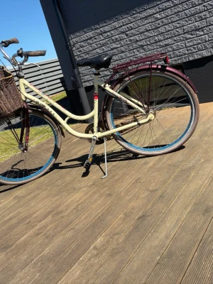 Crescent damcykel med korg - Säljer en klassisk Crescent damcykel i beige och vinrött med en praktisk brun flätad cykelkorg framtill. Cykeln har pakethållare, stänkskärmar och blåa detaljer på däcken. Sadel och handtag är svarta. Perfekt för dig som vill ha en retroinspirerad cykel.