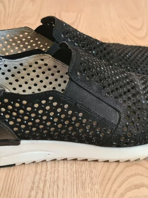 Svarta loafers med hålmönster - Säljer ett par svarta loafers med coolt hålmönster och glittrande detaljer. Skorna har vit sula och elastiska partier på sidorna för enkel påtagning. Materialet ser ut att vara mocka eller liknande textil. Perfekta för dig som vill sticka ut lite extra.