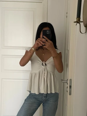 Vit omlottopp med knyt fram - Söt vit topp med omlottkänsla och knytning framtill. Toppen har v-ringning, volangig nederdel och är ärmlös med breda axelband. Perfekt till jeans eller kjol för en fräsch look.
